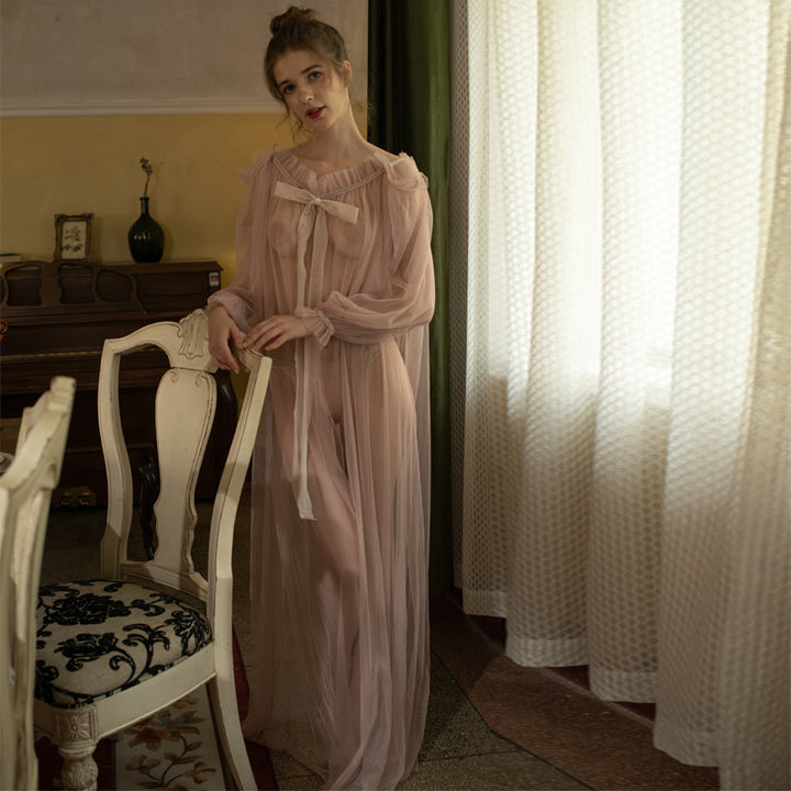 Model wearing a sheer blush-colored lingerie gown with a bow detail, standing beside an ornate chair in a softly lit room with vintage decor.