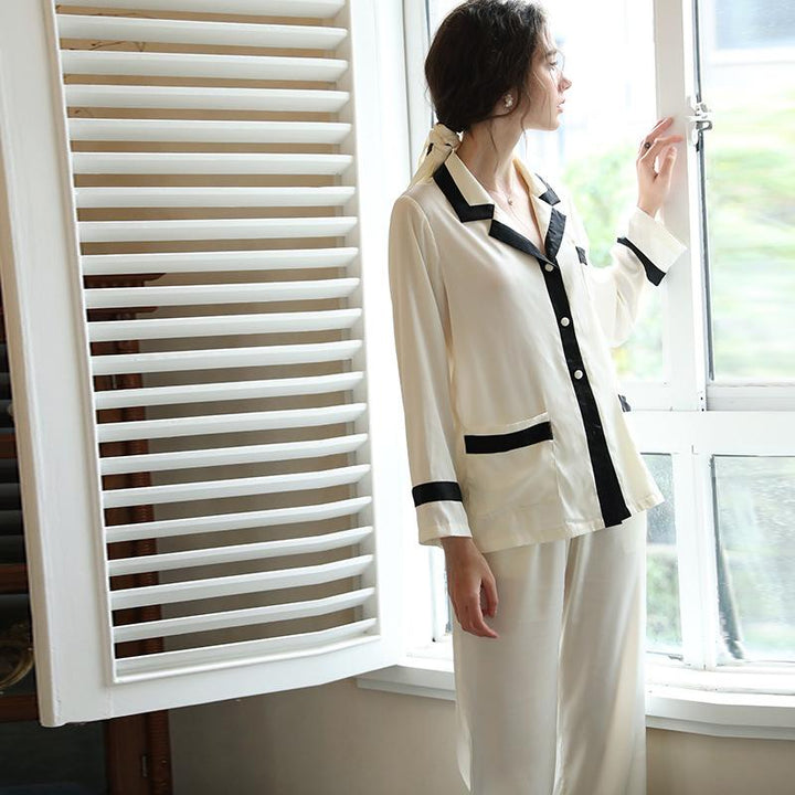 Elegant women's silk pajama set in cream with black trim, featuring a button-up top and matching trousers, styled for comfort and sophistication.