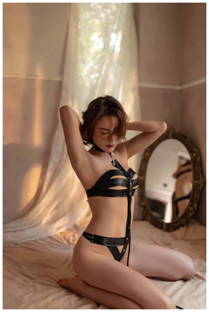 Black faux leather lingerie set featuring cut-out detail and adjustable straps, styled on a model in a softly lit bedroom.