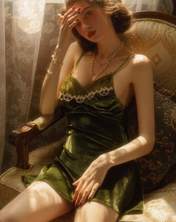 Elegant green velvet lingerie slip dress with lace detailing, modeled by a woman seated on an ornate chair, softly illuminated by natural light.