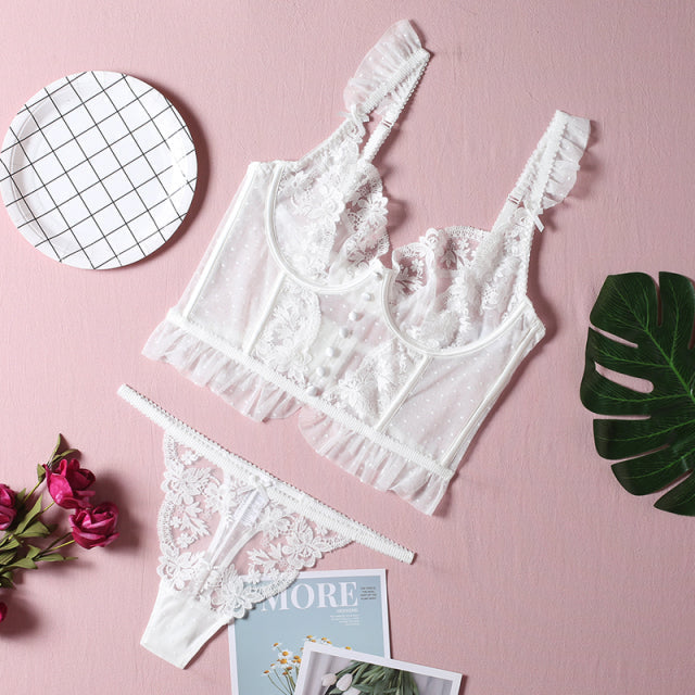 White lace lingerie set featuring a sheer bralette with frill detailing and matching thong, styled with floral accents on a pink background.