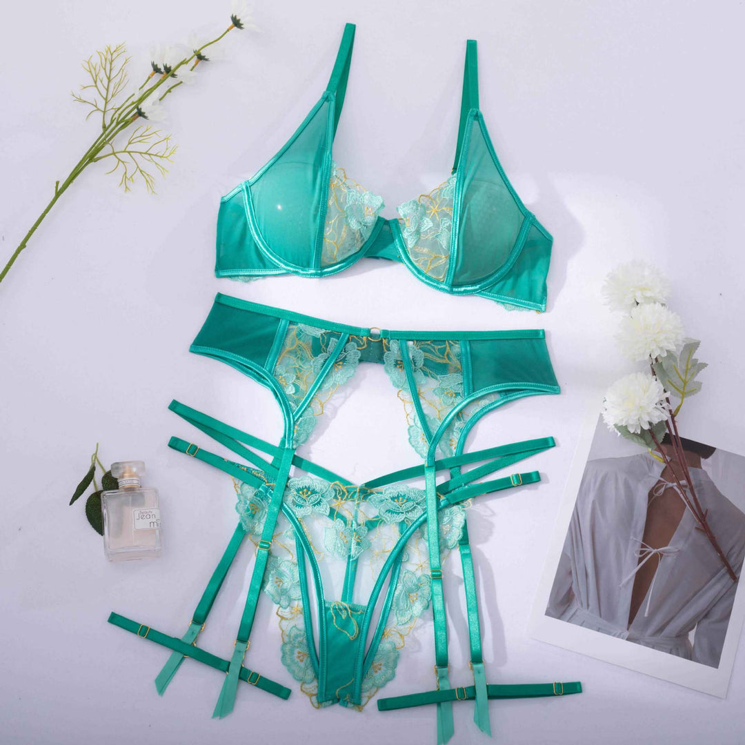 Elegant teal lingerie set featuring a sheer bra with lace detailing and a matching panty with harness-style straps for a sensual look, styled with floral accents and a fragrance bottle.