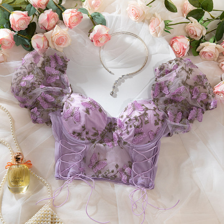 Romantic lilac lingerie top featuring intricate butterfly embroidery, paired with delicate puff sleeves, surrounded by soft fabric, pink roses, a pearl necklace, and