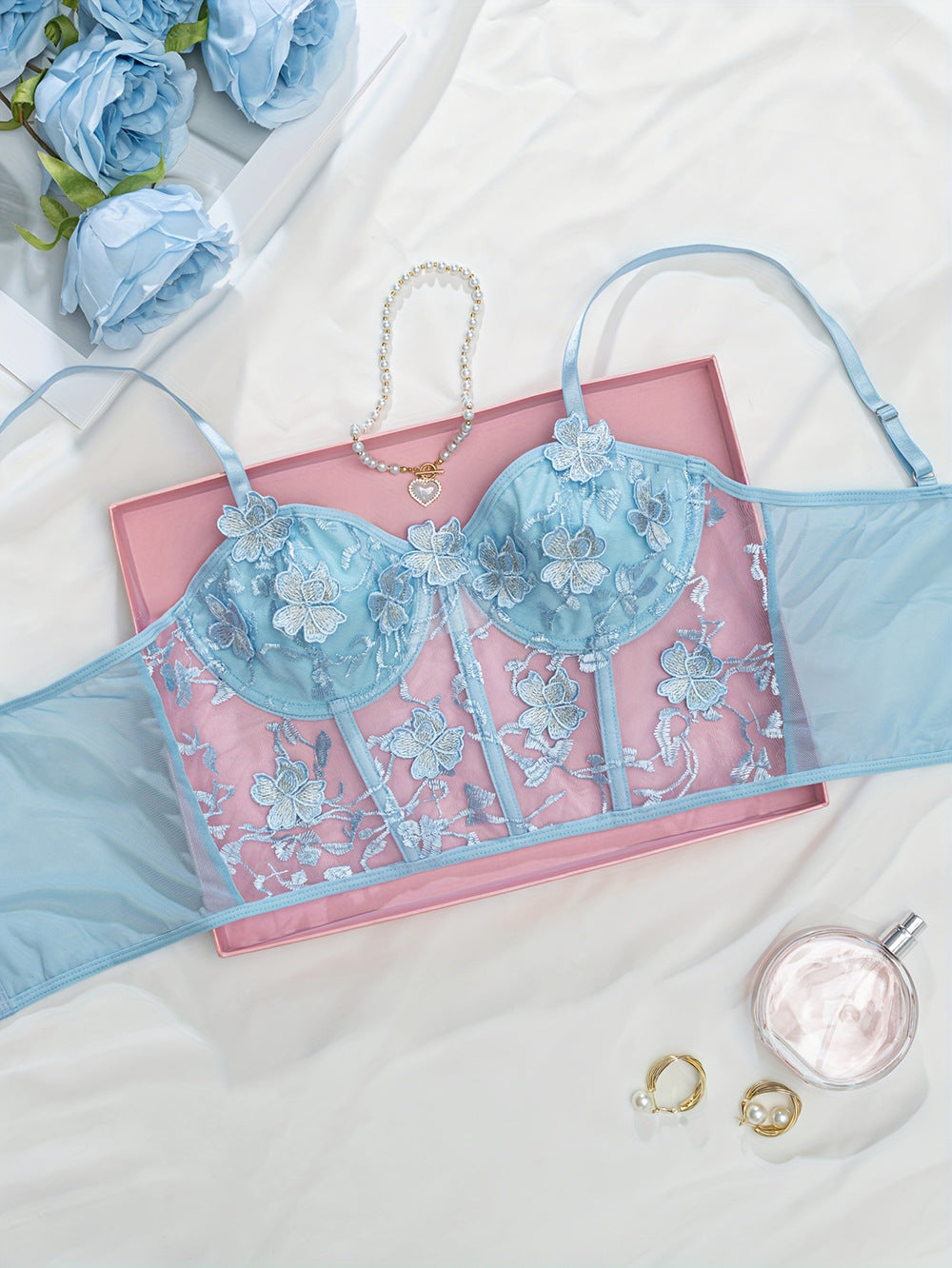 Beautiful blue lace lingerie set featuring floral embroidery, presented on a soft white fabric background alongside pink packaging, a delicate pearl necklace, and elegant jewelry pieces,