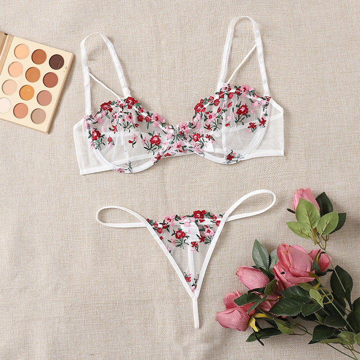 Sheer floral lace lingerie set featuring a soft-cup bra adorned with intricate red and pink embroidery, paired with matching thong panties, all arranged on a