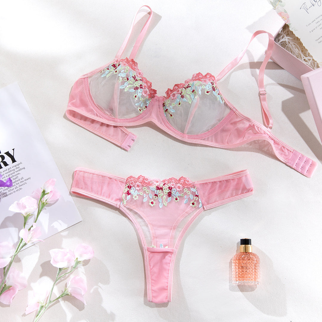 Pink lingerie set featuring a delicate bralette with floral embroidered lace details and matching thong. The ensemble is styled on a light background alongside a floral arrangement and