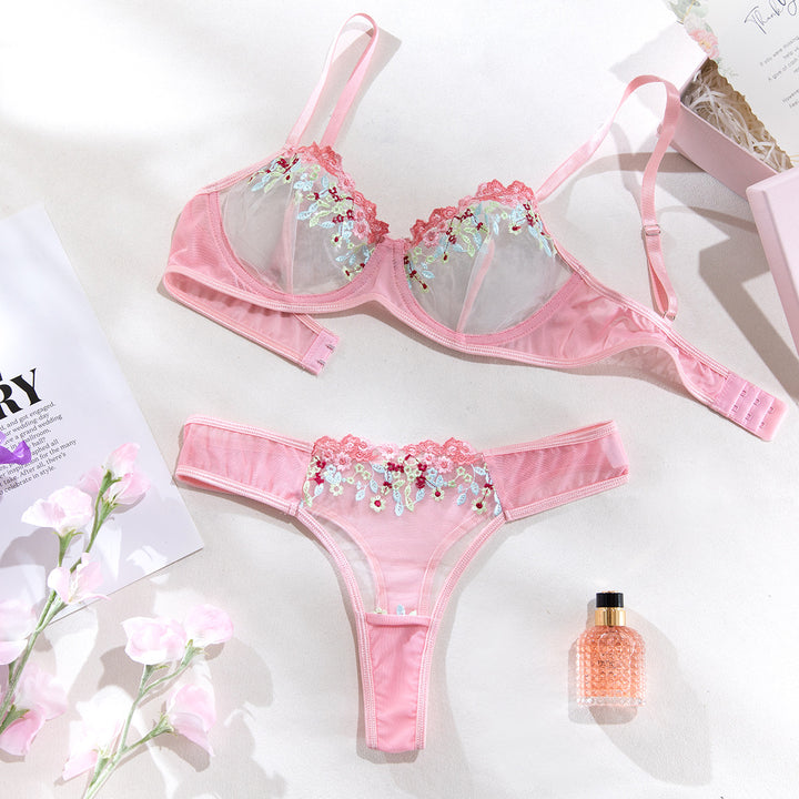 Pink lingerie set featuring a delicate bralette with floral embroidered lace details and matching thong. The ensemble is styled on a light background alongside a floral arrangement and