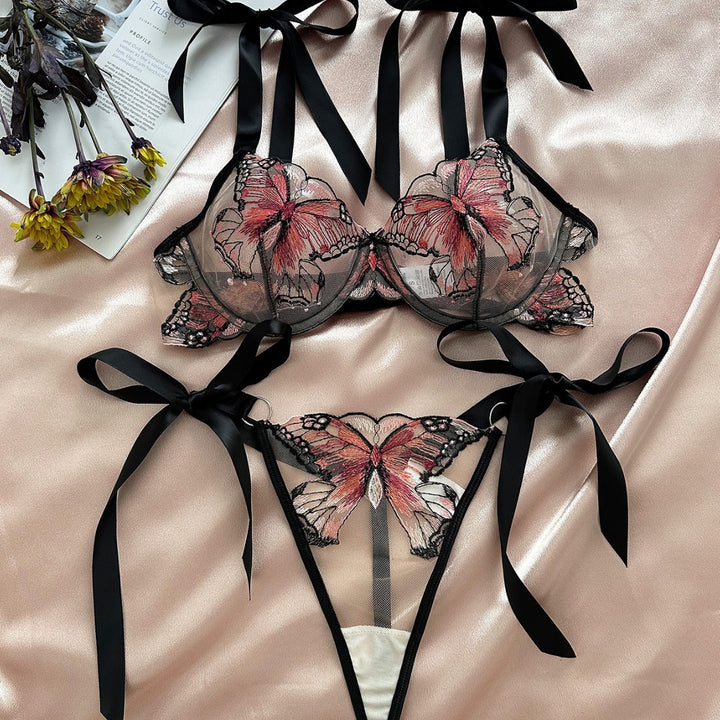 Lingerie set featuring a black mesh bra and thong adorned with vibrant butterfly embroidery, complemented by satin ribbons, displayed on a silky background with floral accents