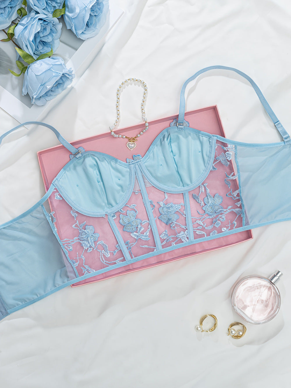 Blue embroidered lingerie top showcased on a delicate pink box, surrounded by silk fabric, faux blue roses, a pearl bracelet with a heart charm, and elegant