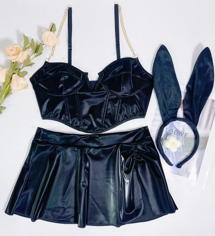 Black satin lingerie set featuring a structured bralette with chain straps, a flared skater skirt, and a bunny ear headband, styled with soft flowers and fashion magazine backdrop.