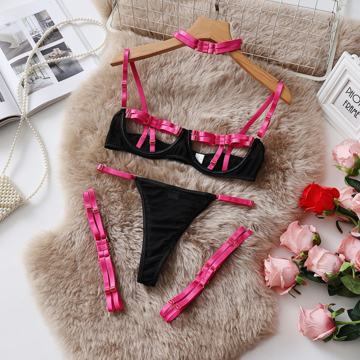 Black lingerie set with pink accents, featuring a bralette with decorative straps, matching thong, and adjustable straps, displayed on a soft fur background with roses and a stylish clutch.