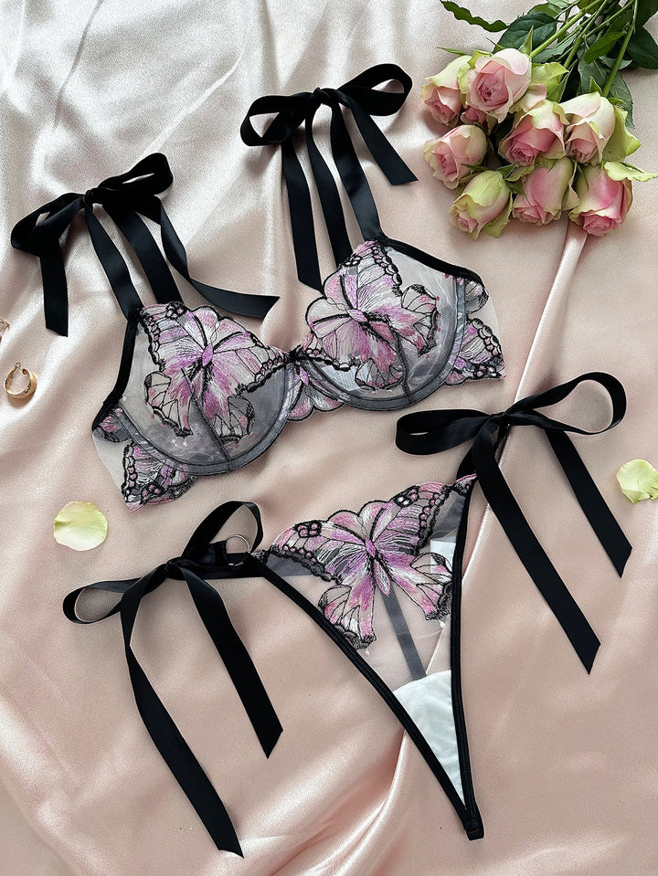 Elegant black lingerie set featuring floral lace embroidery with pink butterfly designs, adorned with feminine black satin ribbons. The set includes a bra and matching thong, styled