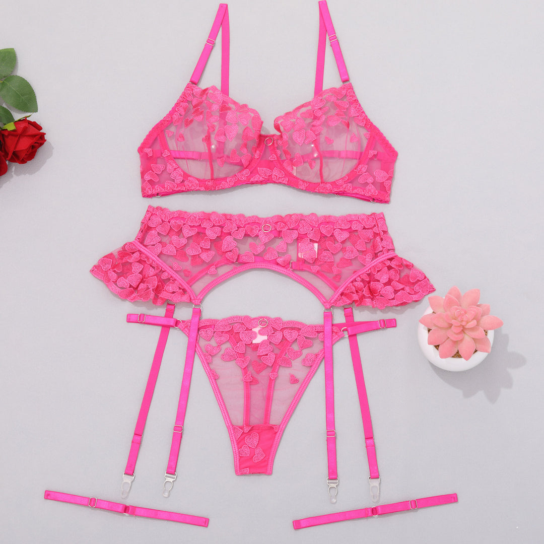 Pink lace lingerie set with heart patterns, featuring a bra, garter belt, and thong, arranged elegantly on a light background with decorative plants.