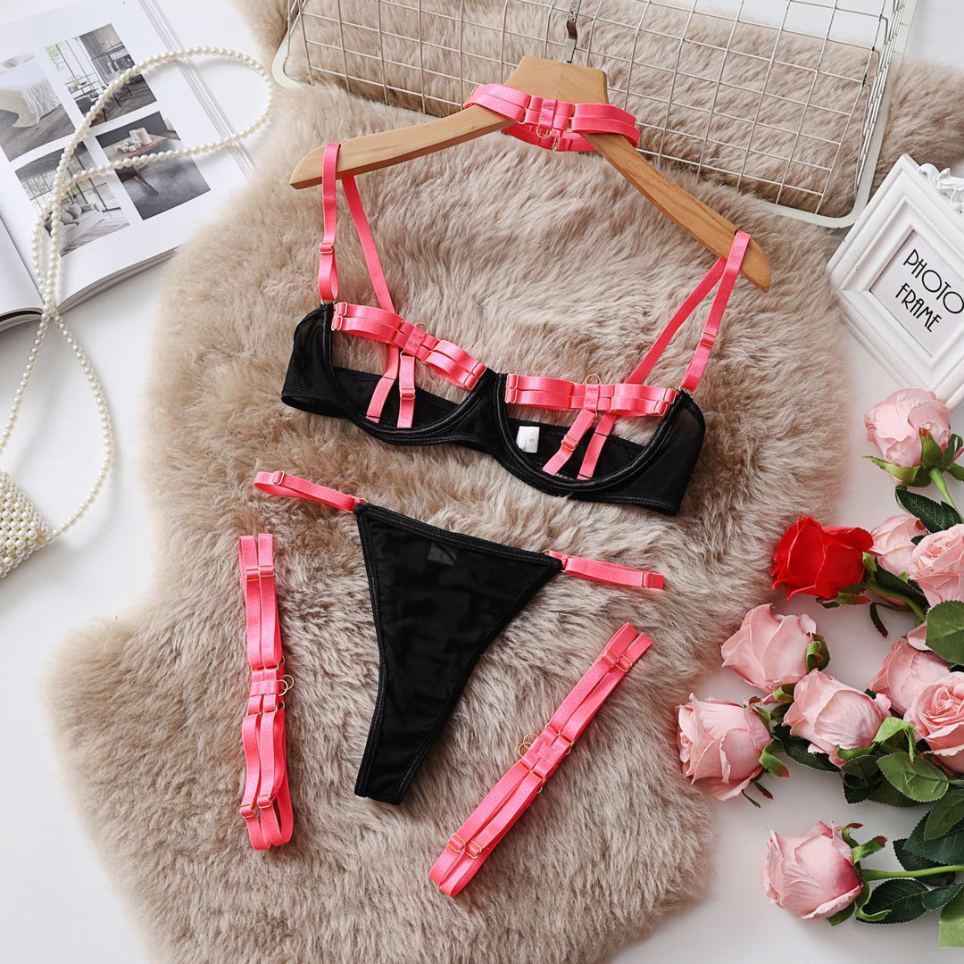 Black lingerie set featuring a bra with pink satin straps and matching thong, styled with decorative harnesses, displayed on a fluffy surface with roses.