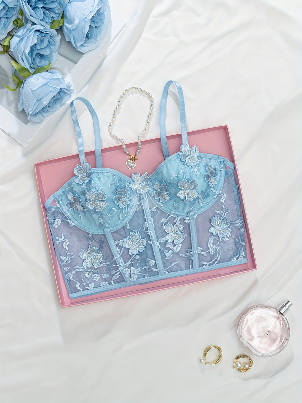 Blue embroidered lace lingerie bralette with floral detailing, presented in a pink gift box, accompanied by a pearl necklace, golden rings, and a bottle of