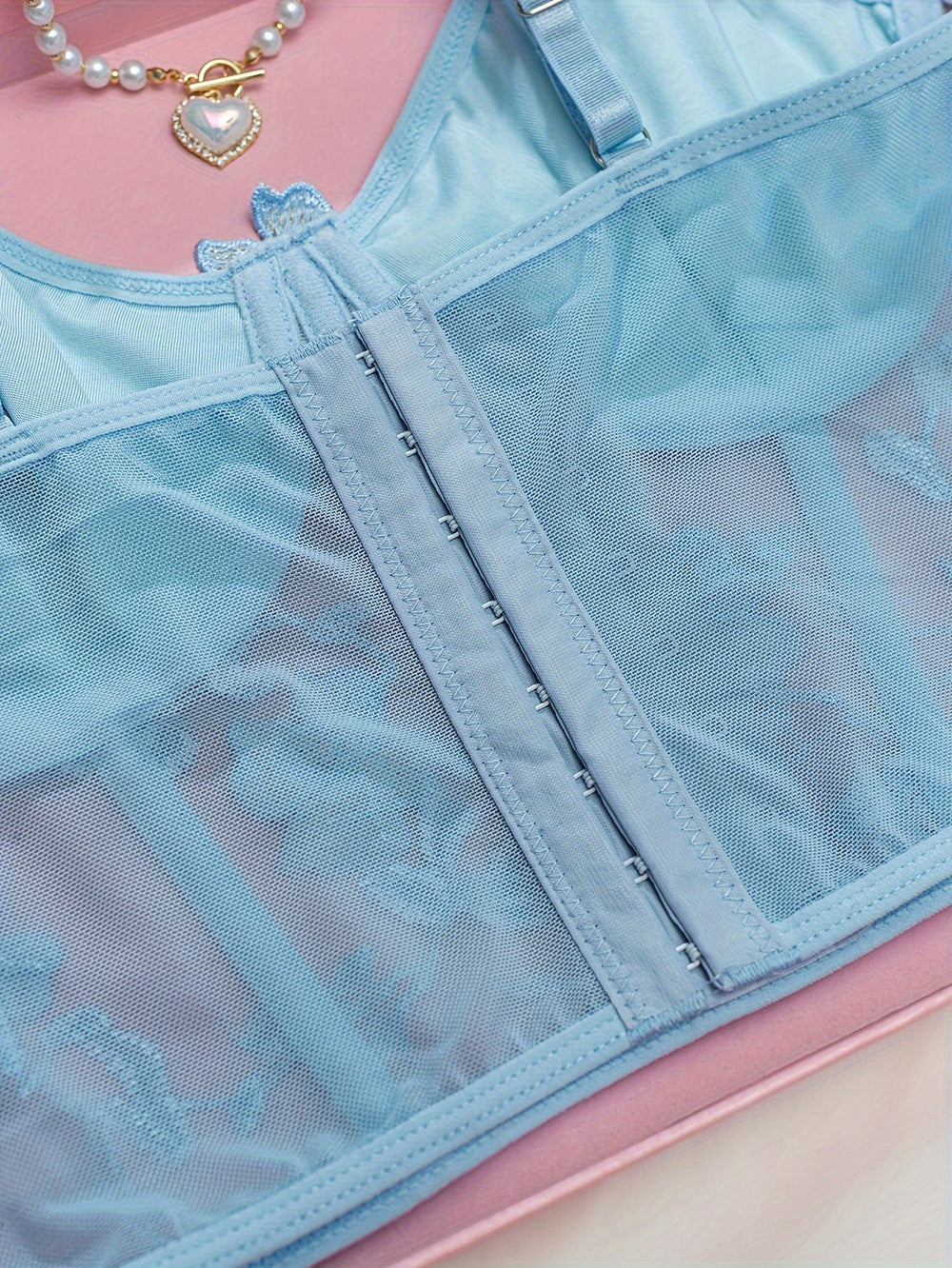 Light blue lingerie with delicate mesh detailing, featuring an intricate hook closure and adjustable straps. Styled on a soft pink backdrop, accented by a pearl necklace with