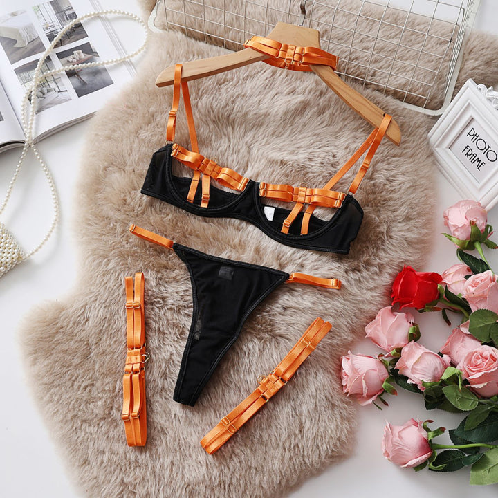 Black lingerie set with orange straps featuring a bra, thong, and strap accessories on a soft beige fur surface with pink roses and a decorative frame in the background.