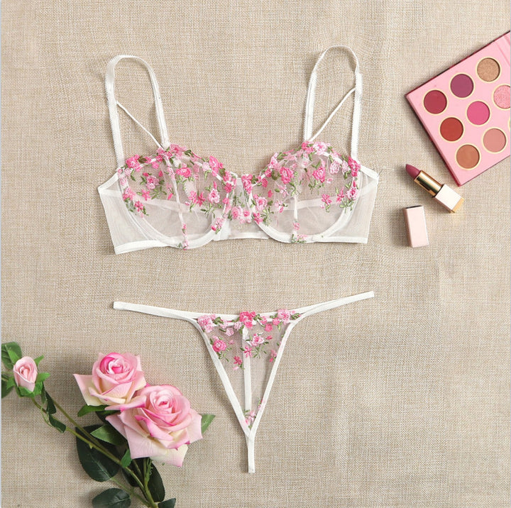 Luxe floral lace lingerie set featuring a delicate sheer bralette adorned with pink embroidered flowers and a matching thong, styled with pink roses and makeup products for