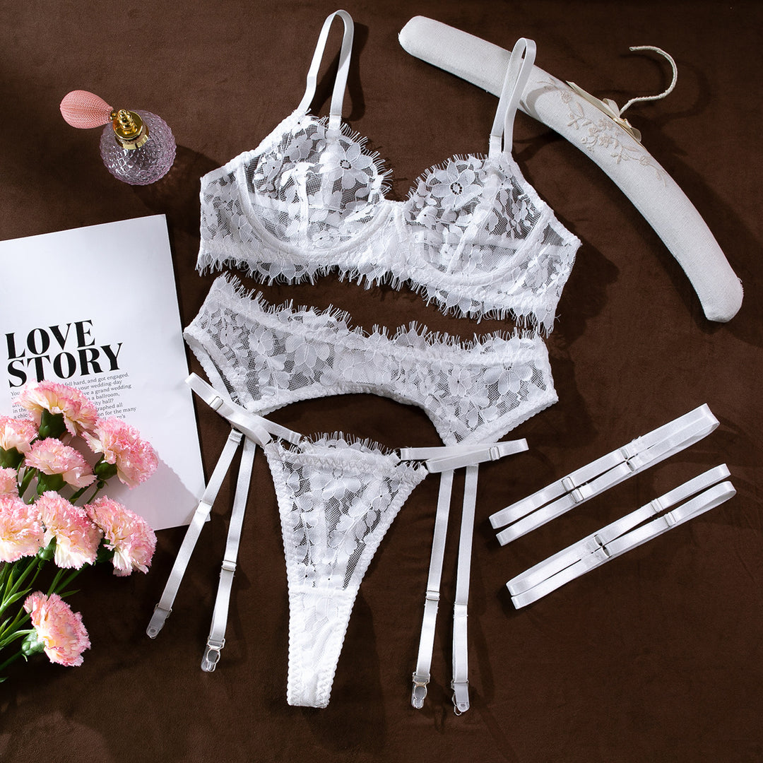 White lace lingerie set featuring a bralette, matching thong, and suspender straps, styled with pink flowers and a magazine on a soft brown background.