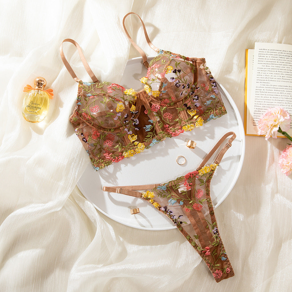 Lingerie set featuring a sheer, embroidered bra and matching thong in earthy tones, adorned with colorful floral designs. Styled with a delicate golden ring and