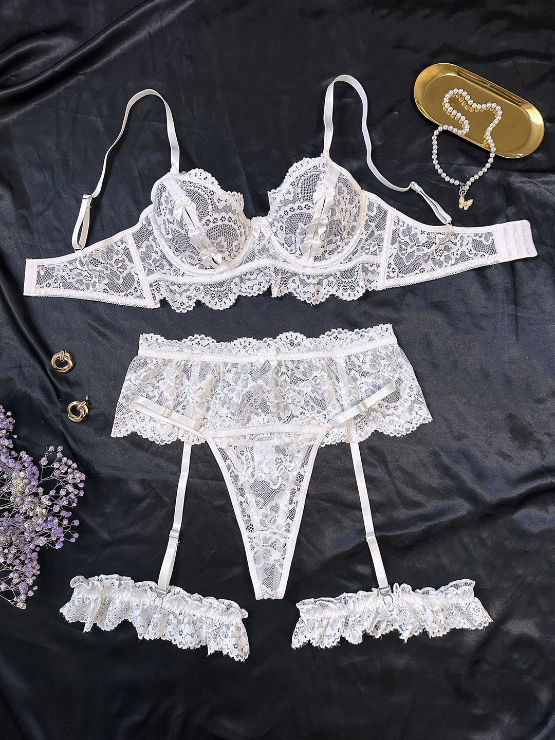 White lace lingerie set featuring a balconette bra, garter belt, and matching thong with delicate ruffle details, styled on a dark satin background.