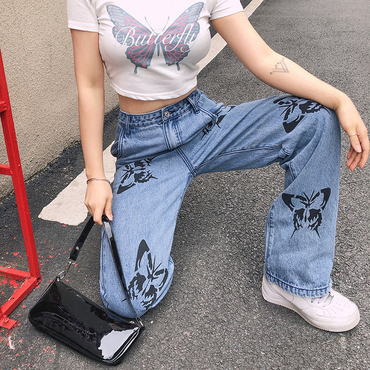 Model wearing a white butterfly graphic crop top with high-waisted blue jeans featuring butterfly prints, paired with a black handbag and white sneakers, posing in an urban setting.