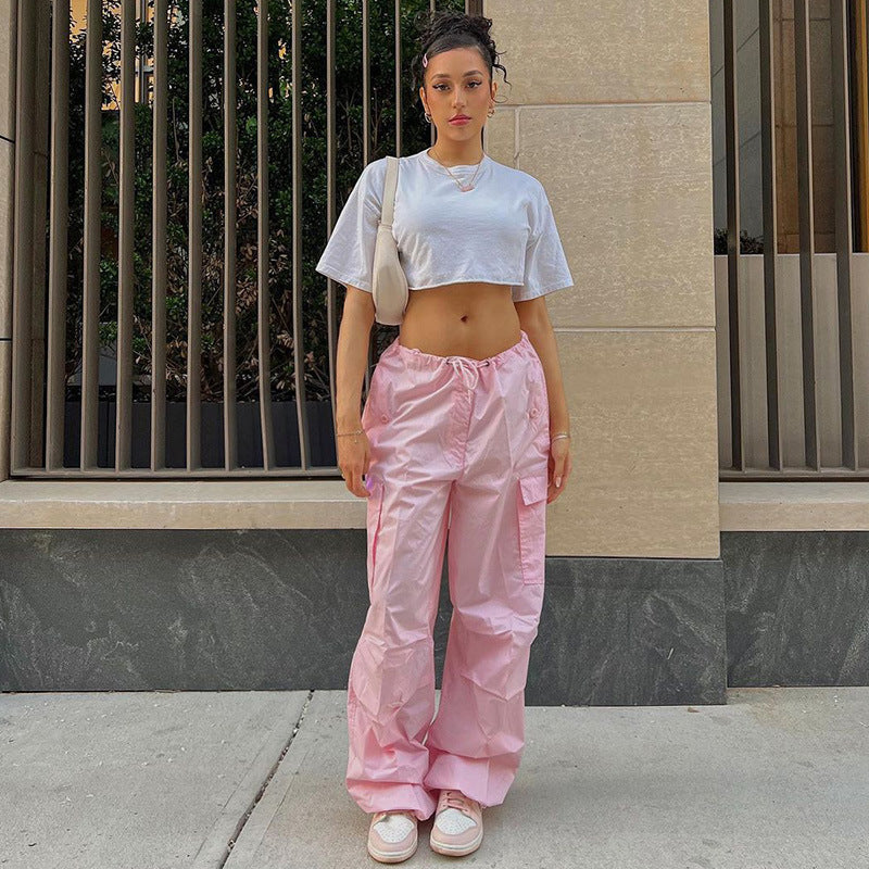 Model wearing a cropped white t-shirt and pink cargo pants, standing against a textured wall with vertical bars, showcasing a trendy streetwear style.