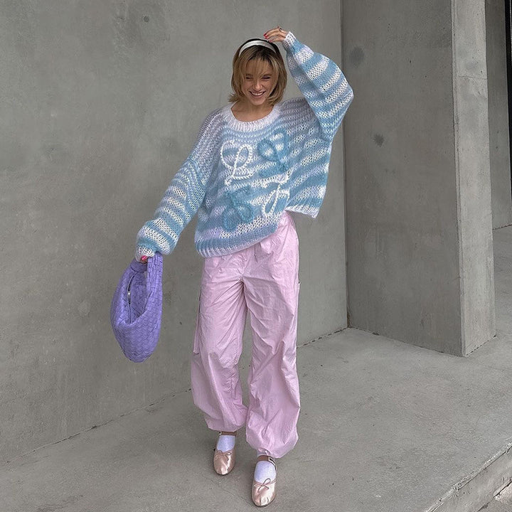 Model wearing a cozy blue and white striped oversized sweater with playful lettering, paired with pastel pink oversized pants, holding a purple bag in a minimalist indoor setting.