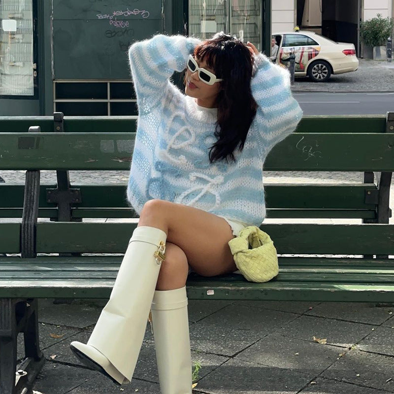 Model wearing a cozy blue striped sweater, white thigh-high boots, and holding a small woven bag while sitting on a bench in an urban setting.