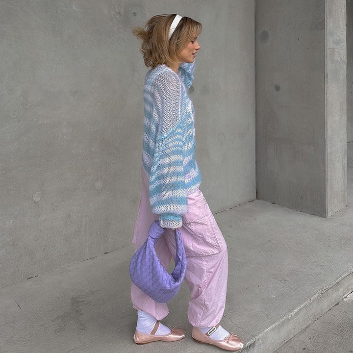 Model wearing a pastel blue and white striped oversized sweater paired with pink trousers, holding a purple woven handbag, styled with a headband and blush flats.