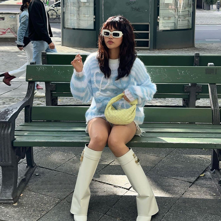 Fashionable woman sitting on a bench wearing a light blue oversized sweater, white high-heeled boots, and holding a yellow handbag, styled with sunglasses in an urban setting.