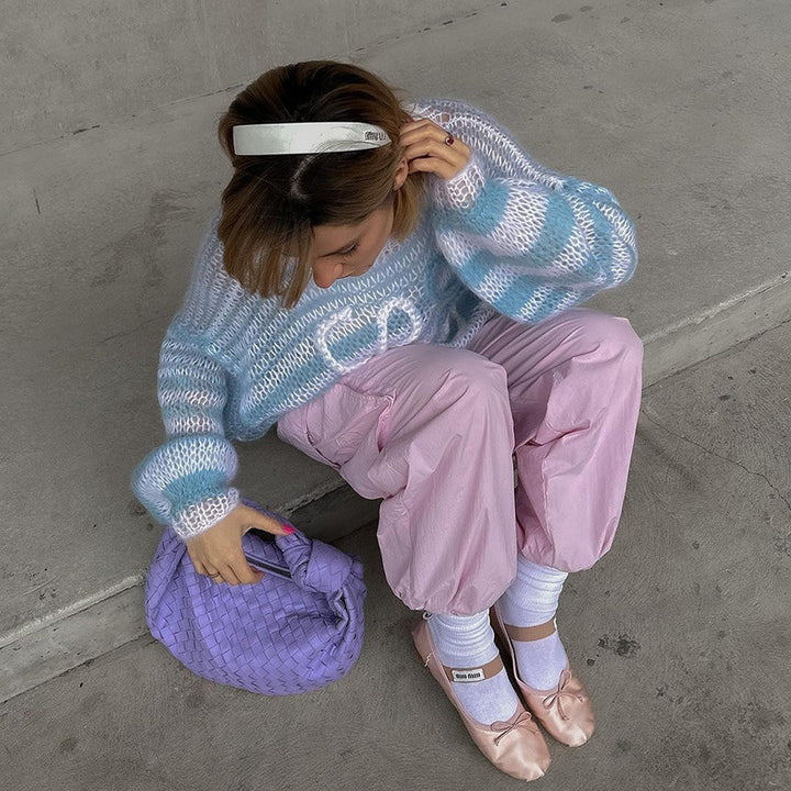 Woman in a cozy blue and white striped sweater, pink baggy pants, and ballet flats, sitting on the ground with a lavender handbag.