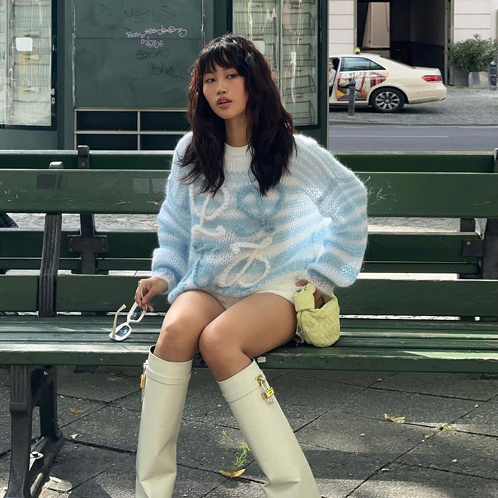 Model wearing a light blue and white oversized knitted sweater with a drawstring, paired with white knee-high boots, sitting on a green bench outdoors.