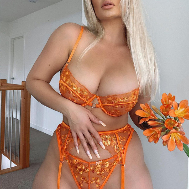 Orange floral lace lingerie set featuring a bra and matching high-waisted panties, model holding a bouquet of orange flowers, stylish indoor setting.