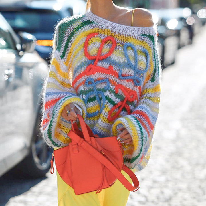 Colorful oversized knit sweater with playful designs, worn casually with bright yellow pants and an orange handbag, in a sunny outdoor setting.