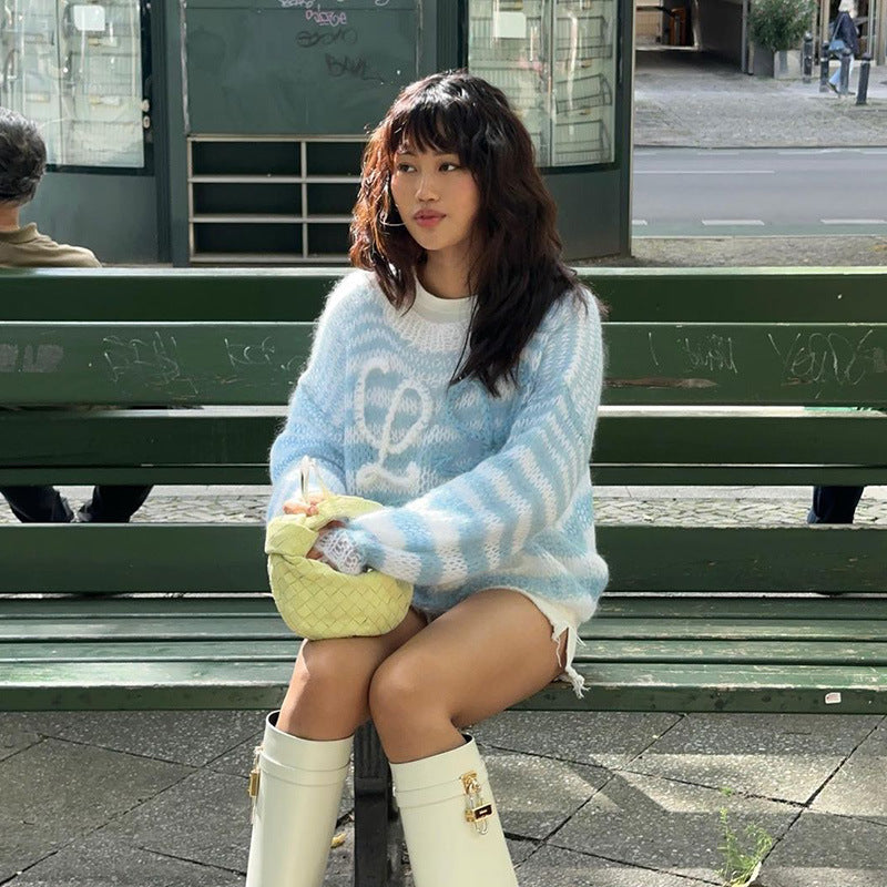 Fashionable model sitting on a park bench wearing a cozy blue and white striped oversized sweater, paired with white knee-high boots and a yellow handbag.