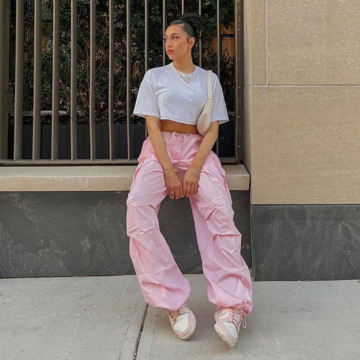 Model wearing a relaxed white crop top and pink cargo pants, sitting stylishly against a modern urban backdrop.