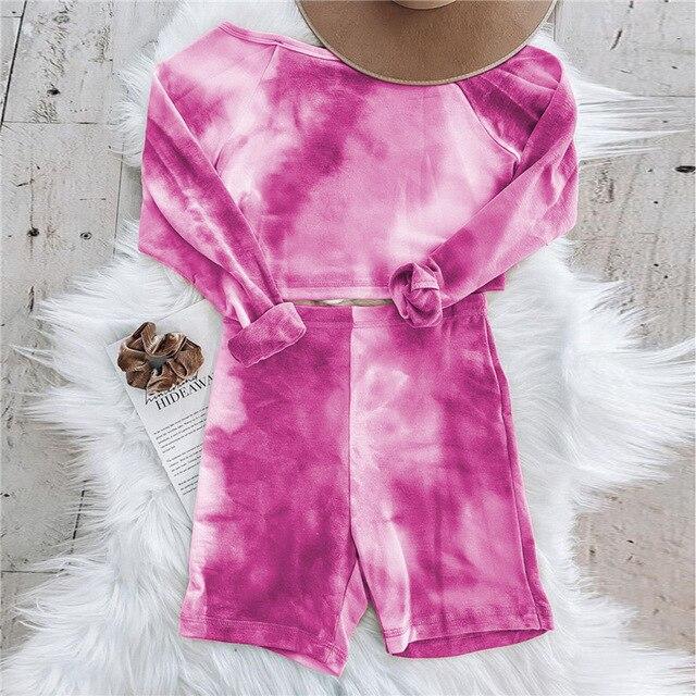 Pink tie-dye long sleeve top and shorts set laid out on a fluffy white surface, accessorized with a brown hat and hair scrunchie.