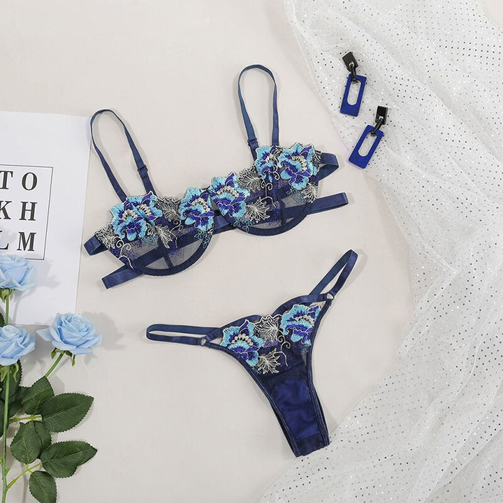 Navy blue floral lace lingerie set featuring a bra with adjustable straps and matching thong, arranged elegantly with decorative accessories and soft roses on a textured surface.